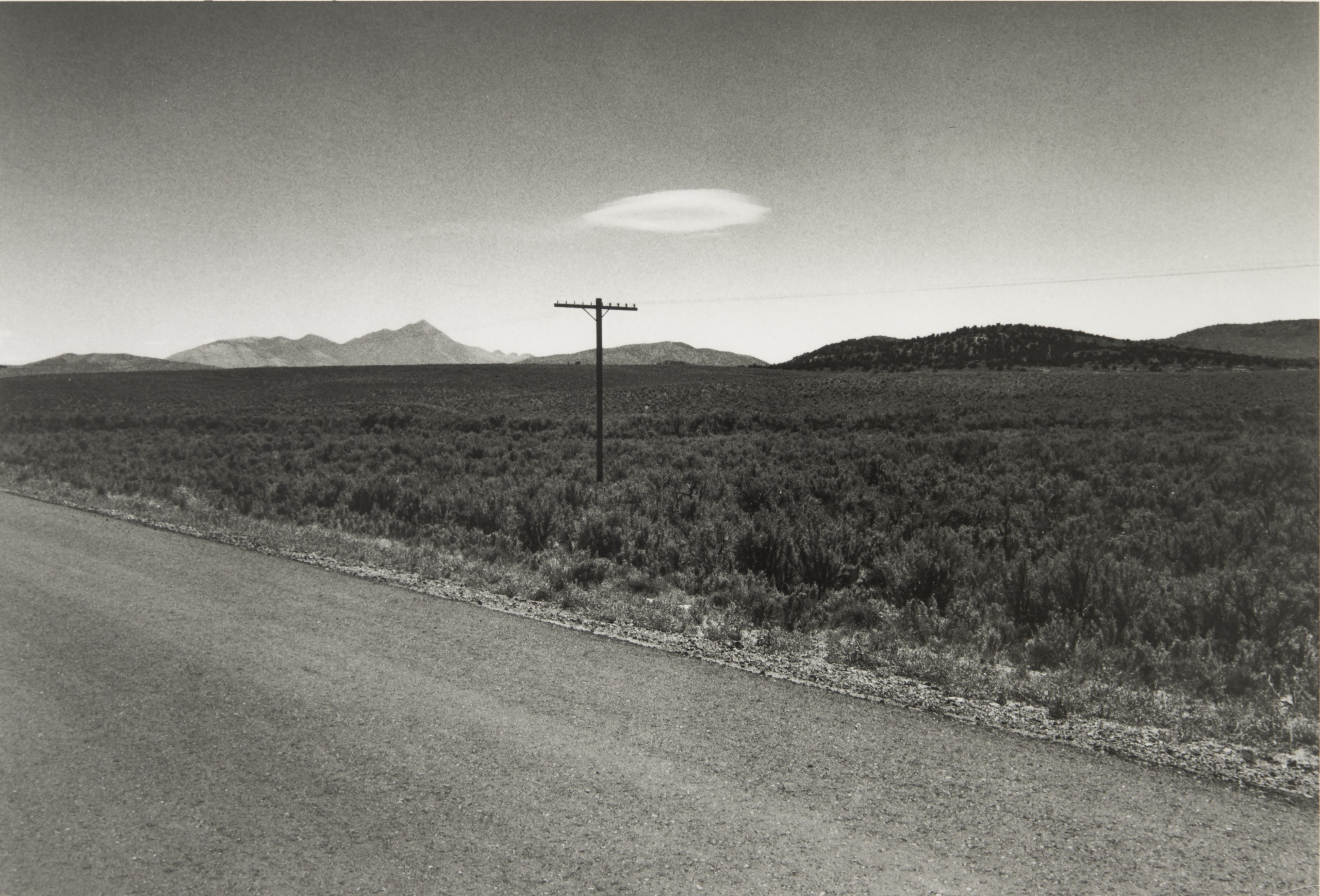 A black and white photograph of a deserted road running past scrubland toward distant mountains, with a telephone pole and a lens-shaped cloud overhead.