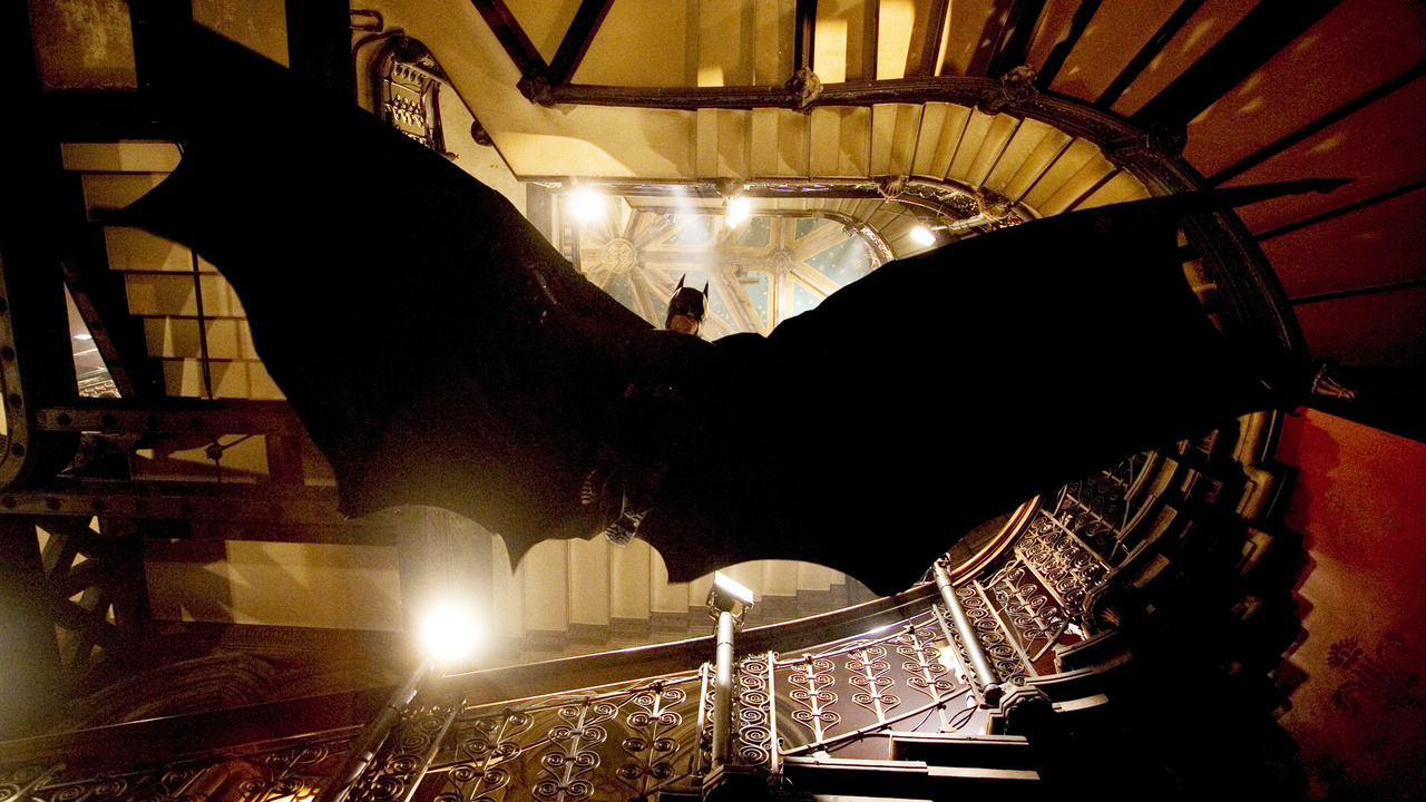 Batman glides down a staircase to evade gunfire in Batman Begins