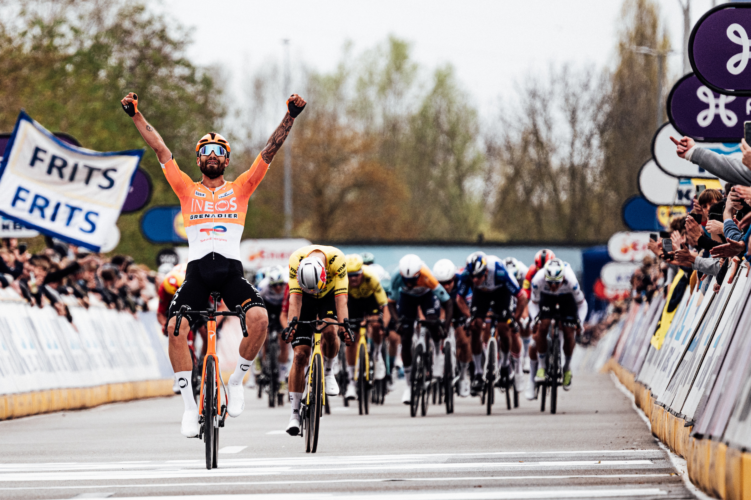 Una foto de Filippo Ganna celebrando la victoria de Dwars en Vlaanderen frente al pelotón de velocidad, con Wout van Aert agachando la cabeza decepcionado. A la derecha de la toma, puedes ver un brazo que pasa por encima de la barrera y le muestra el dedo medio.