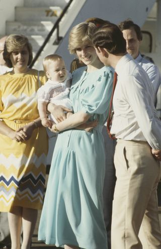 Princess Diana standing outside wearing a blue dress and holding baby Prince William who is looking at Prince Charles