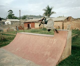 A man doing a one handed hand stand at the top of a skateboard ramp with his skateboard on his feet in the air above him.