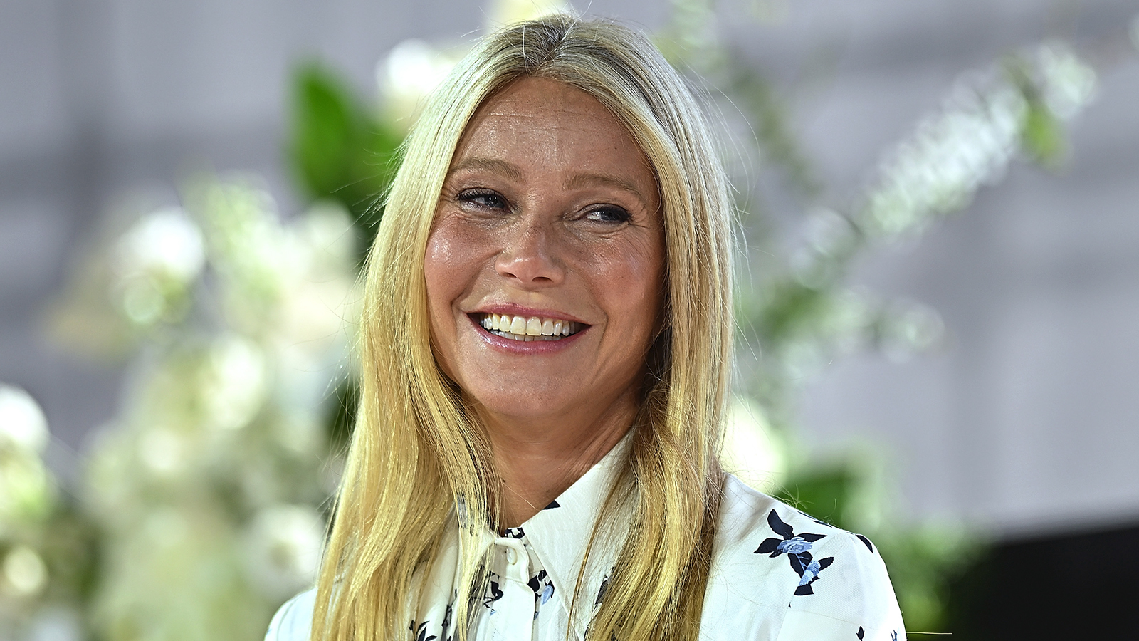 Headshot of Gwyneth Paltrow speaking at Forbes Power Women&#039;s Summit, wearing white blouse with foliage visible in background