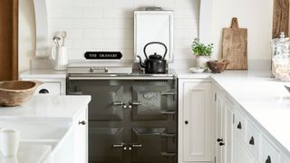 A traditional kitchen features a dark grey range cooker framed by white cabinetry and subway tiles. A black kettle rests on the hob. Wooden boards, ceramics, and simple greenery add texture, creating a calm, pared-back space with classic farmhouse character.