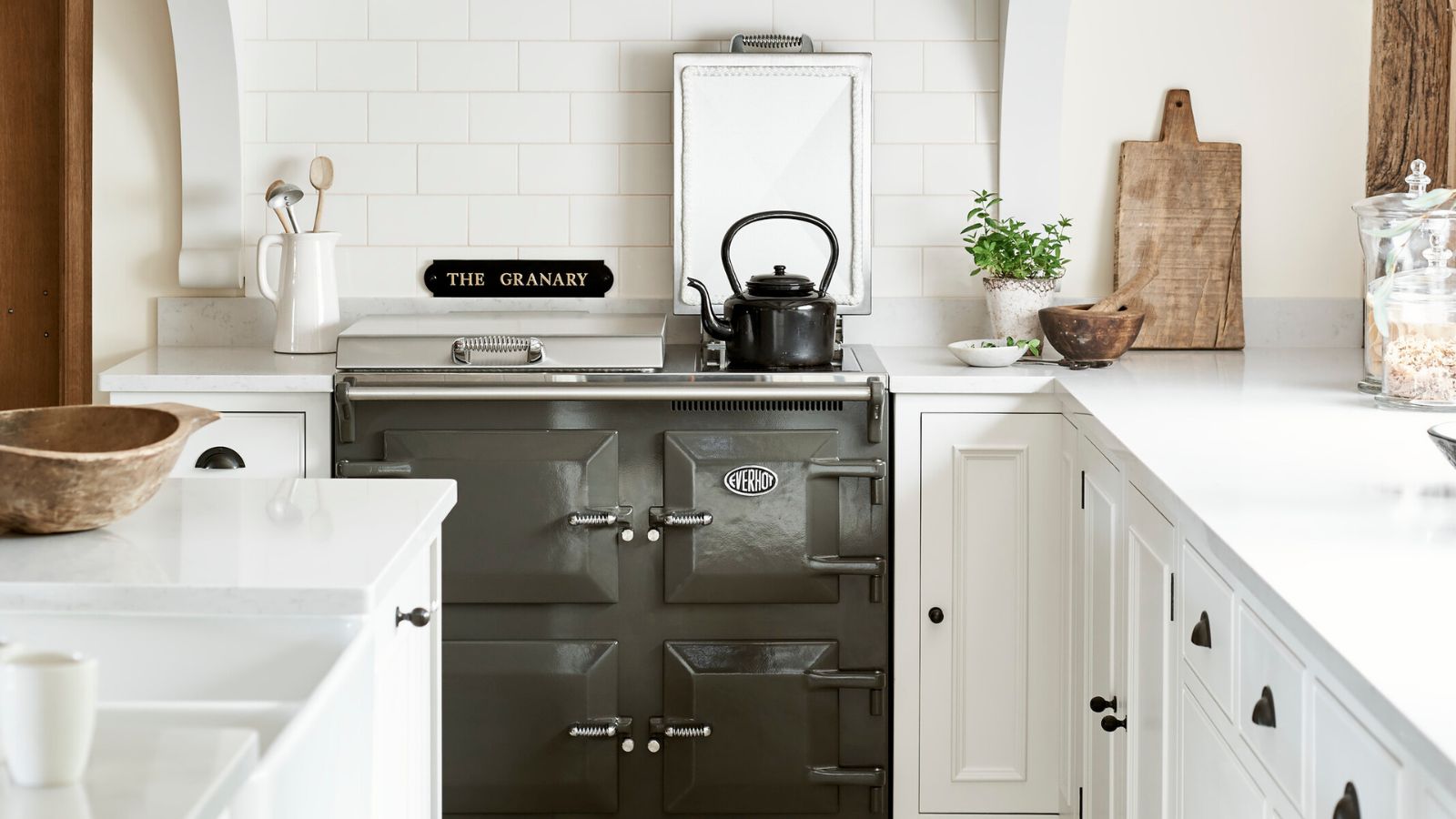 A traditional kitchen features a dark grey range cooker framed by white cabinetry and subway tiles. A black kettle rests on the hob. Wooden boards, ceramics, and simple greenery add texture, creating a calm, pared-back space with classic farmhouse character.