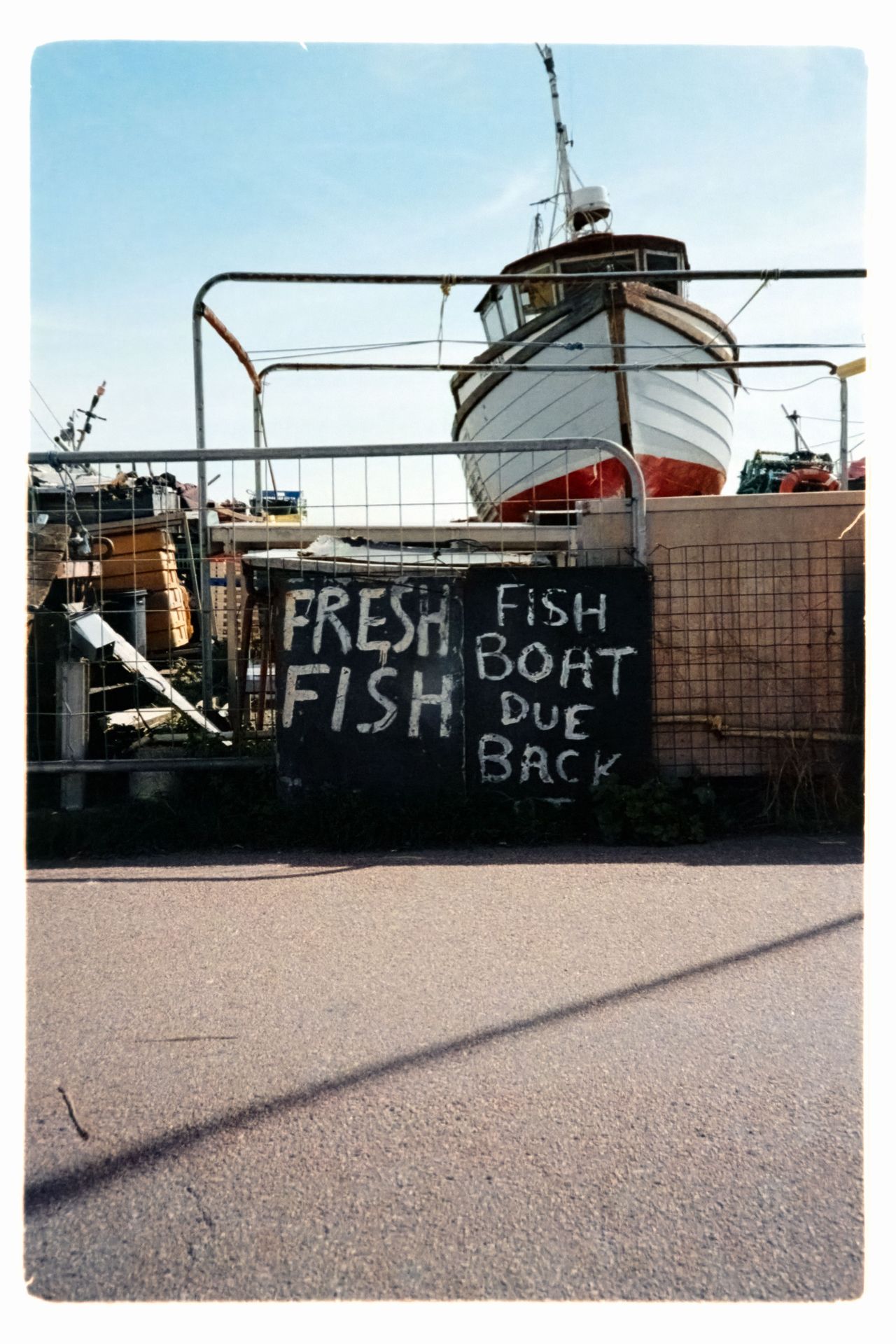 Scanned film image of a fishing boat behind railings, with a hand-painted sign reading &ldquo;Fresh Fish, Fish Boat Due Back.&rdquo;