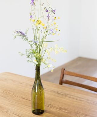 A green glass wine bottle with yellow and purple wildflowers on it, on a light wood countertop with a dark wooden chair underneath it