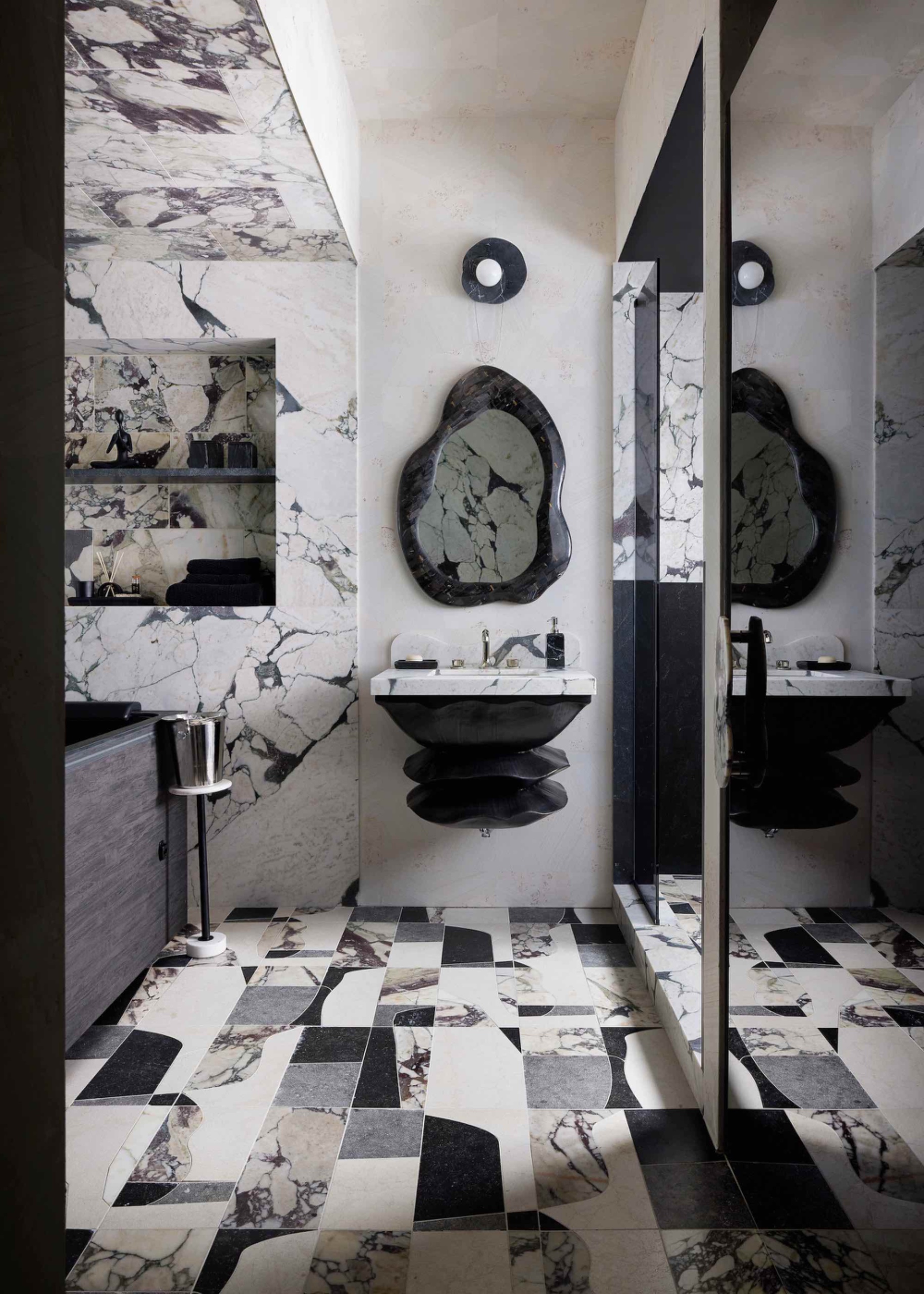 Bathroom with heavily veined marble walls, oversized abstract stone mosaic flooring, a sculptural dark vanity and an irregularly shaped mirror.