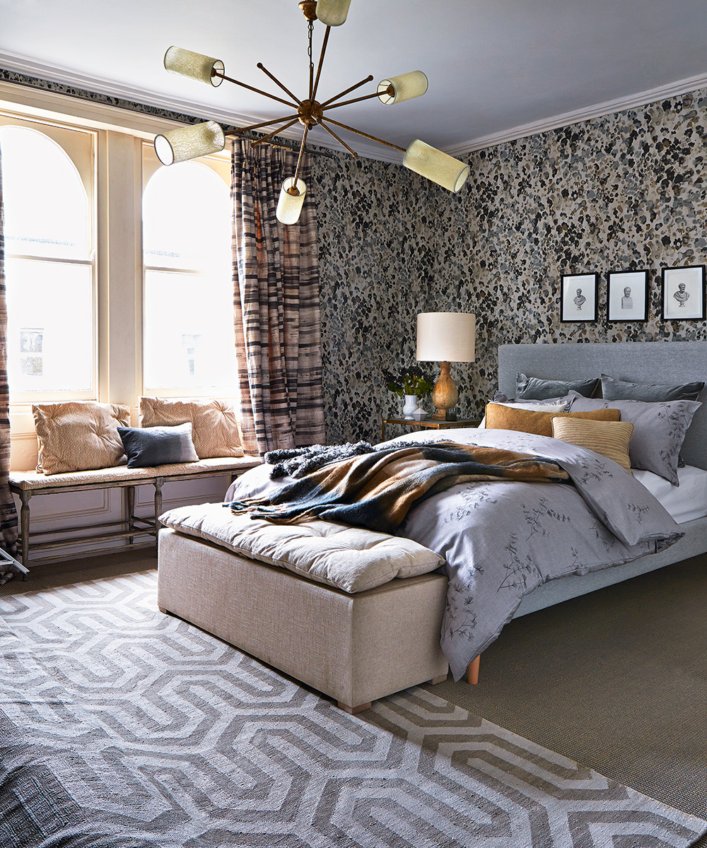 A gray bed with yellow cushions in front of mottled brown and beige wallpaper with a yellow and cream bedside lamp and a pendant ceiling light