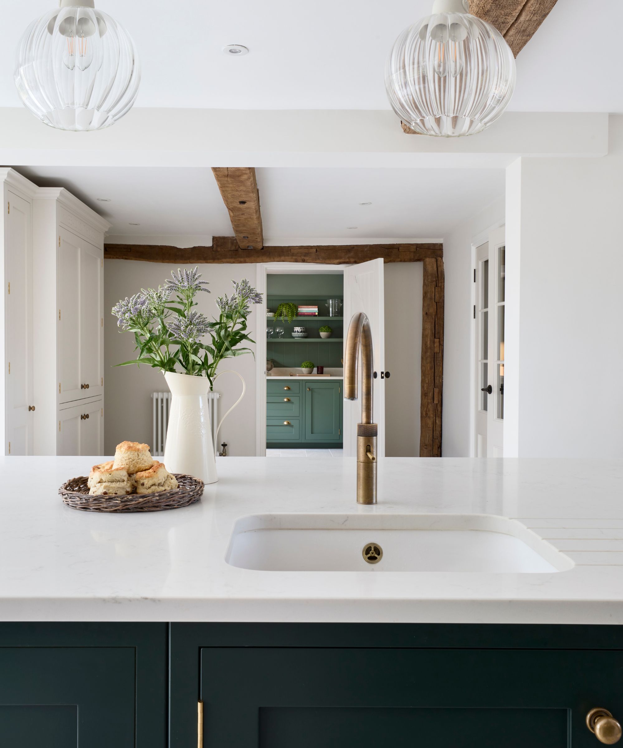 A country-style kitchen with deep green cabinetry, a white quartz worktop, and a brass tap. Rustic beams, soft lighting, and fresh scones add warmth and charm.