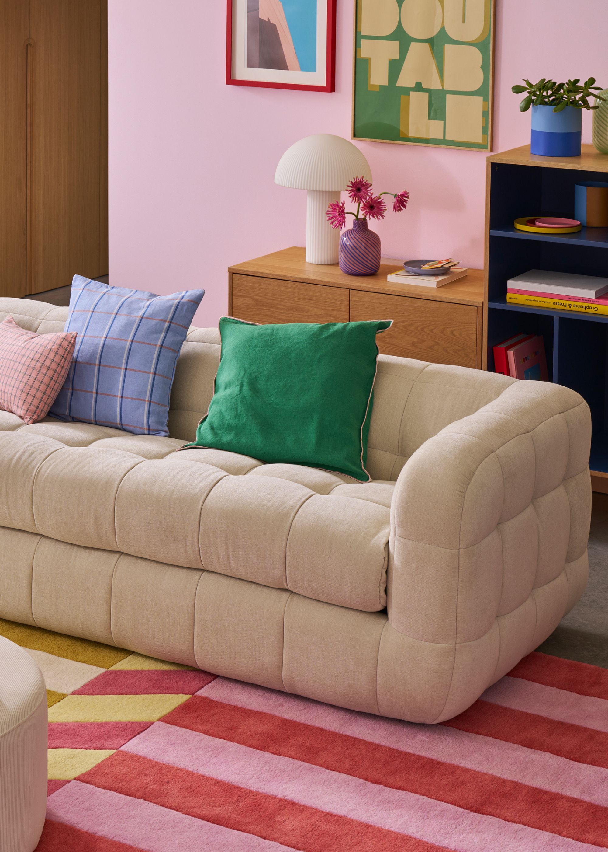 A colorful living room with a pink and red striped rug, pink walls, and a modern cream sofa with green, blue, and pink accent cushions.