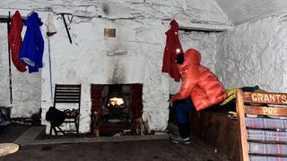 A man sits in a bothy wearing an orange The North Face Summit Series Pumori Down Parka