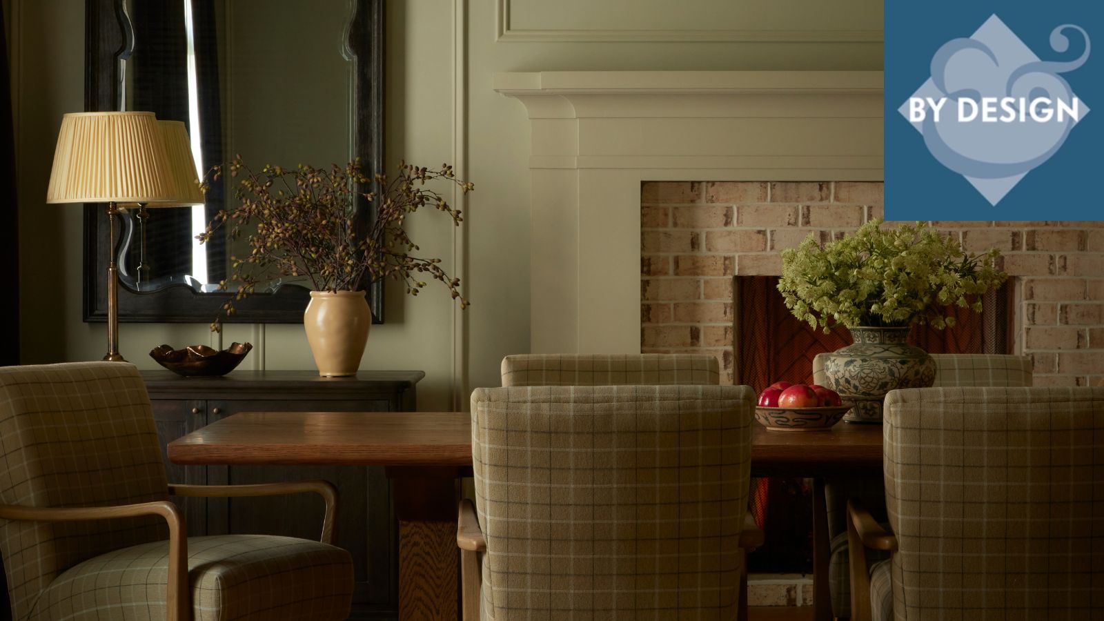Cozy dining room with dark wooden rectangular dining table, upholstered dining chairs, vase of flowers on the table. Brick fireplace, and sideboard with lamp and vase in background.