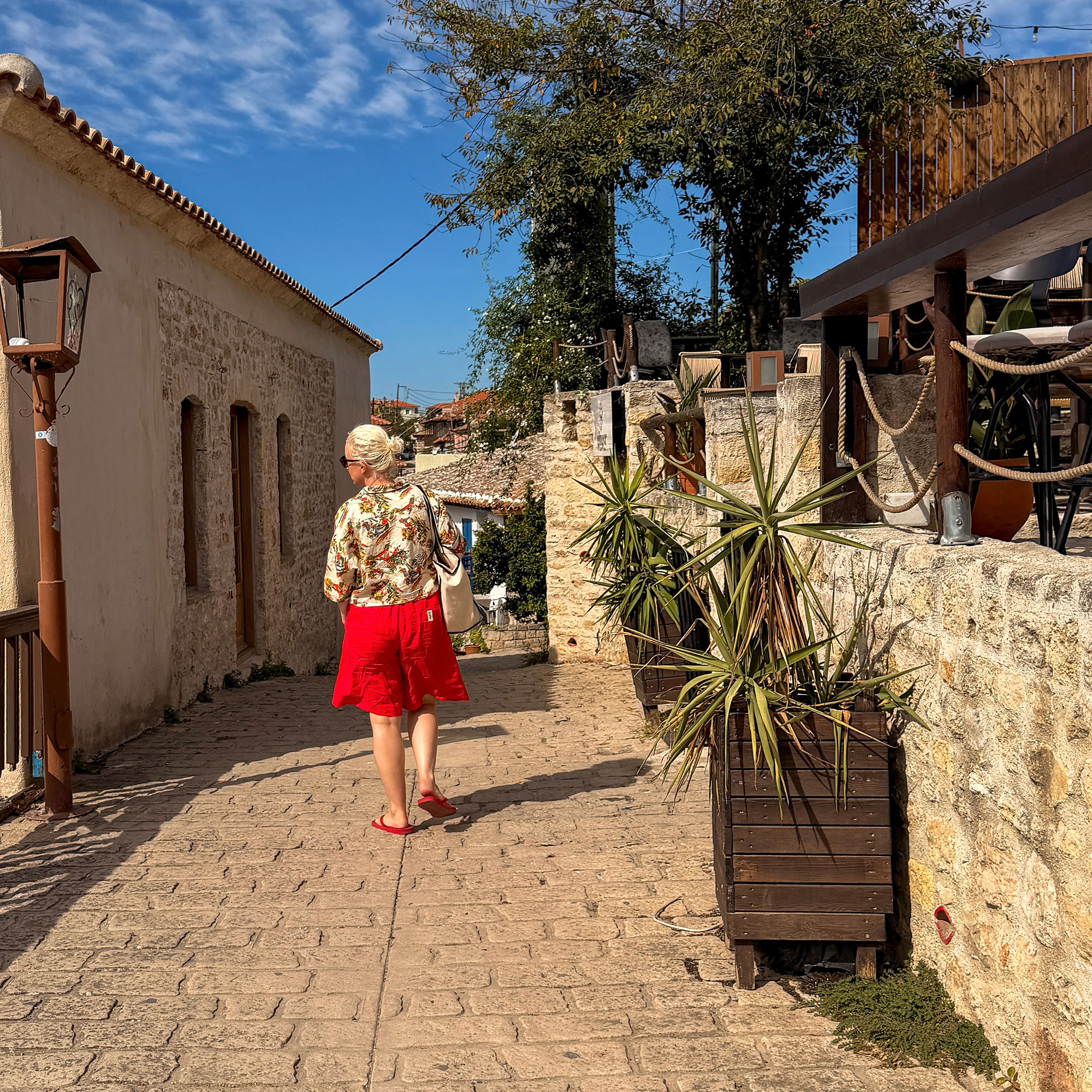 Woman in floral top and long red shorts walking down street with stone walls