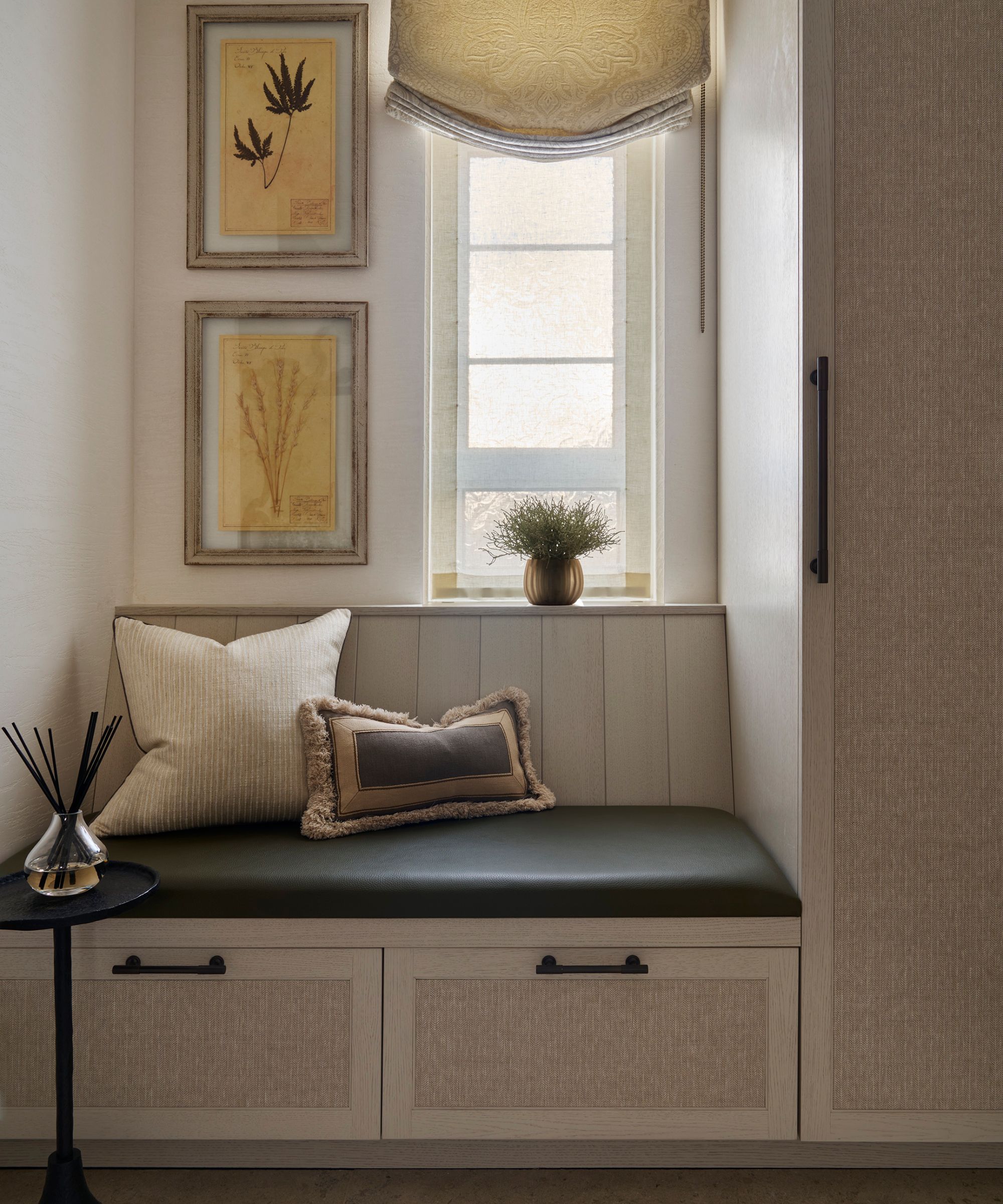 a small storage area with a bench and drawers with a padded cushion, throw pillows, and botanical artwork