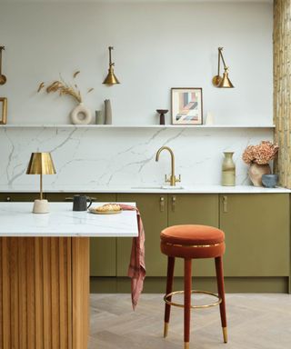 brushed gold metal wall lights with extendable arms above white shelf and kitchen worktop with green cabinet doors, white worktop, small gold table lamp on kitchen island and burnt orange velvet barstool next to island