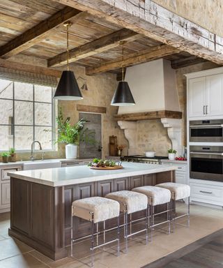 Rustic, country kitchen with exposed woodwork and stone walls, large, dark wooden kitchen island with white marble top, large chimney breast above central stove, two black cone pendants hanging above island