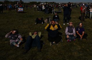 people sit on grass and are wearing solar eclipse viewing glasses.