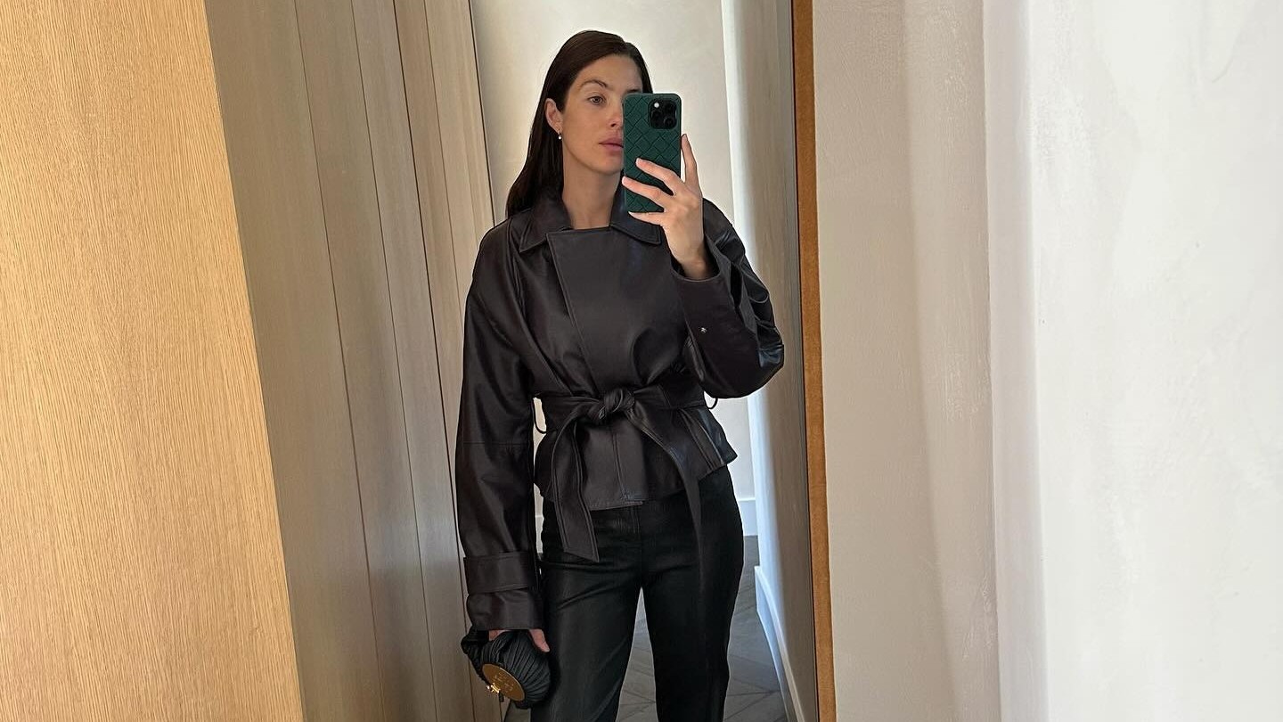 Woman wears black cigarette pants, a leather wrap coat and square toe flats. 