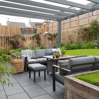 Dining area on a paved patio, with grey outdoor sofas and a matching dining table positioned under a grey pergola
