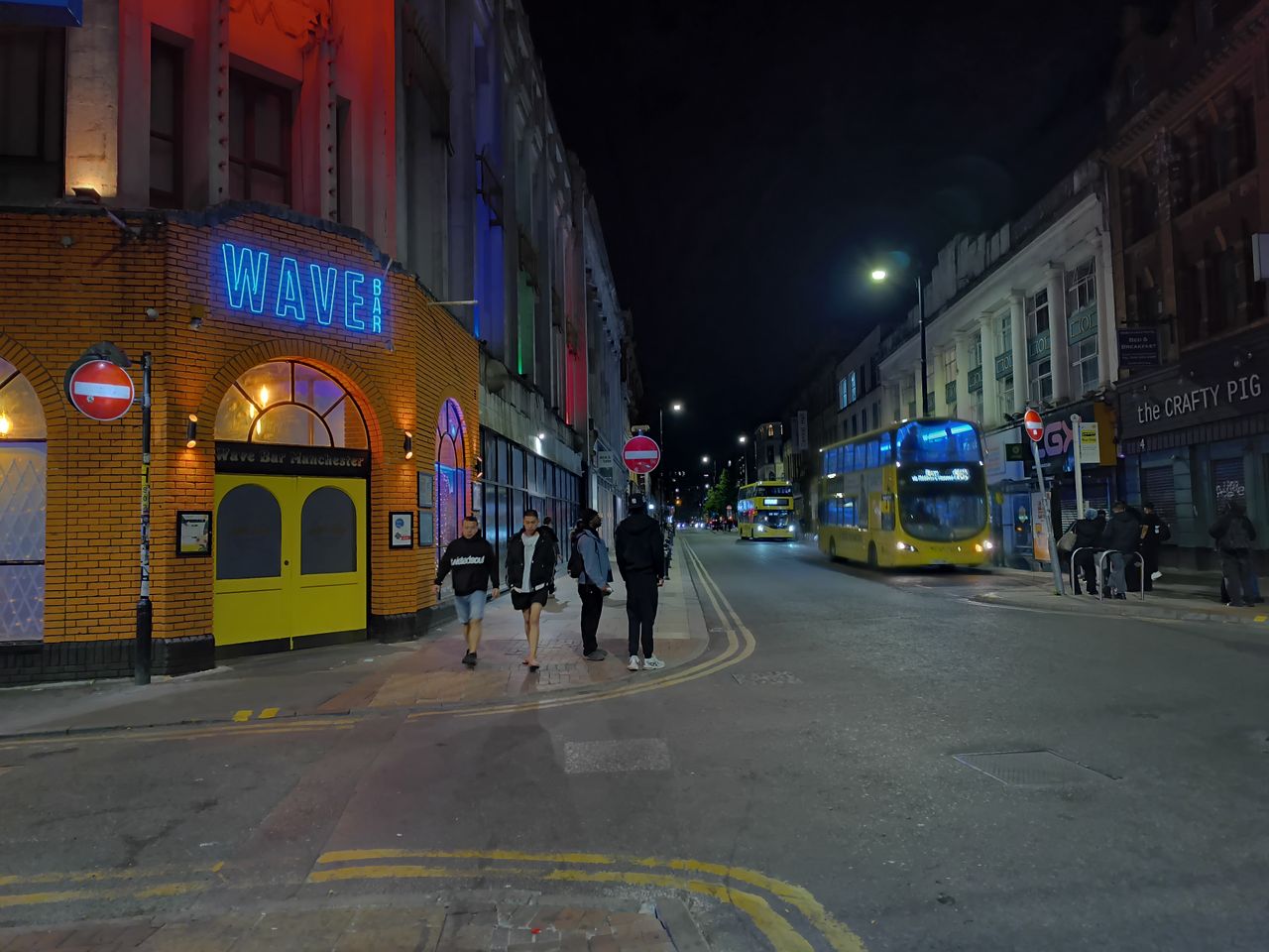 A nighttime street view of WAVE Bar Manchester written in distinctive blue neon signage mounted on an orange brick facade with yellow doors.