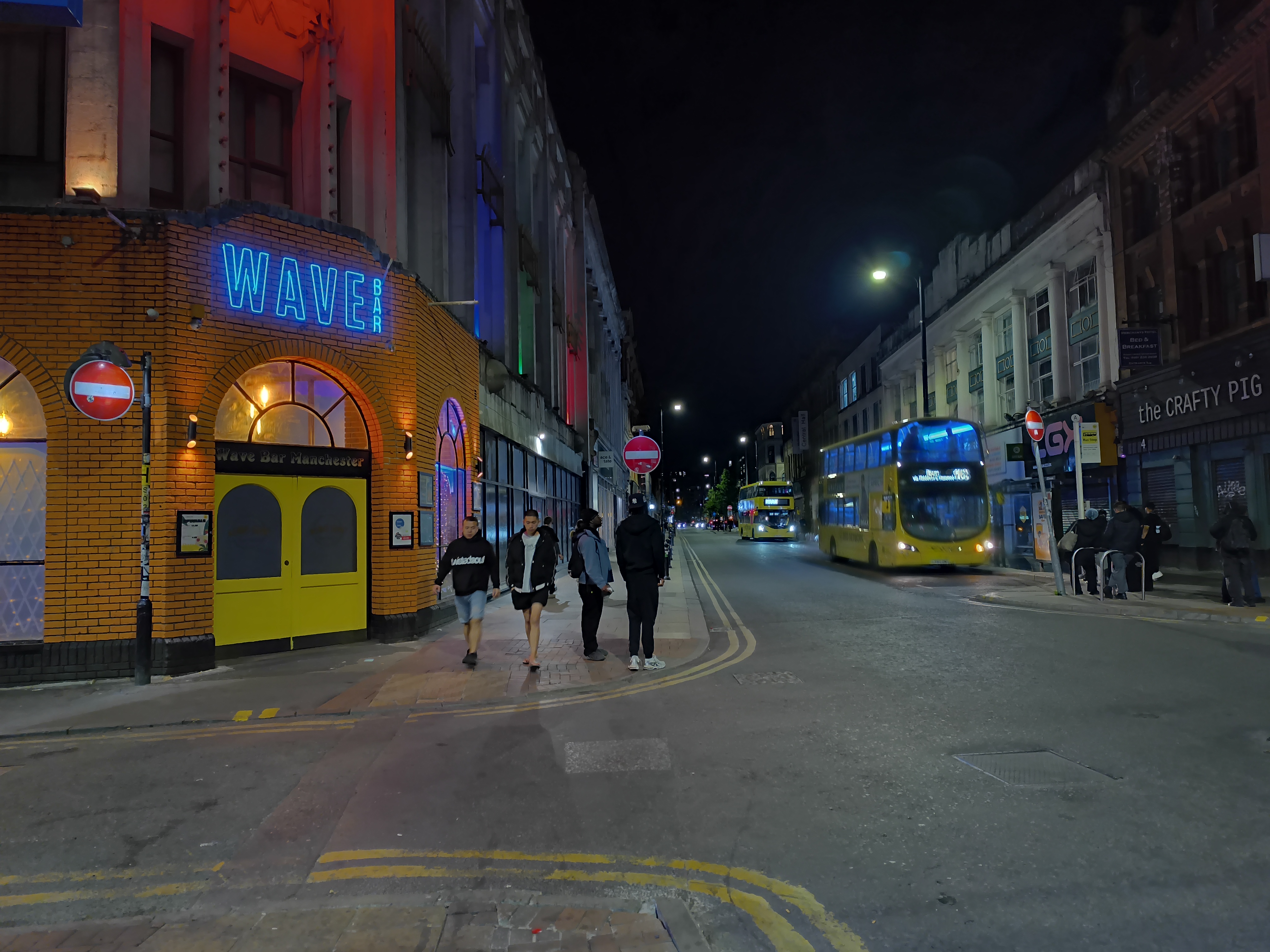 A nighttime street view of WAVE Bar Manchester written in distinctive blue neon signage mounted on an orange brick facade with yellow doors.