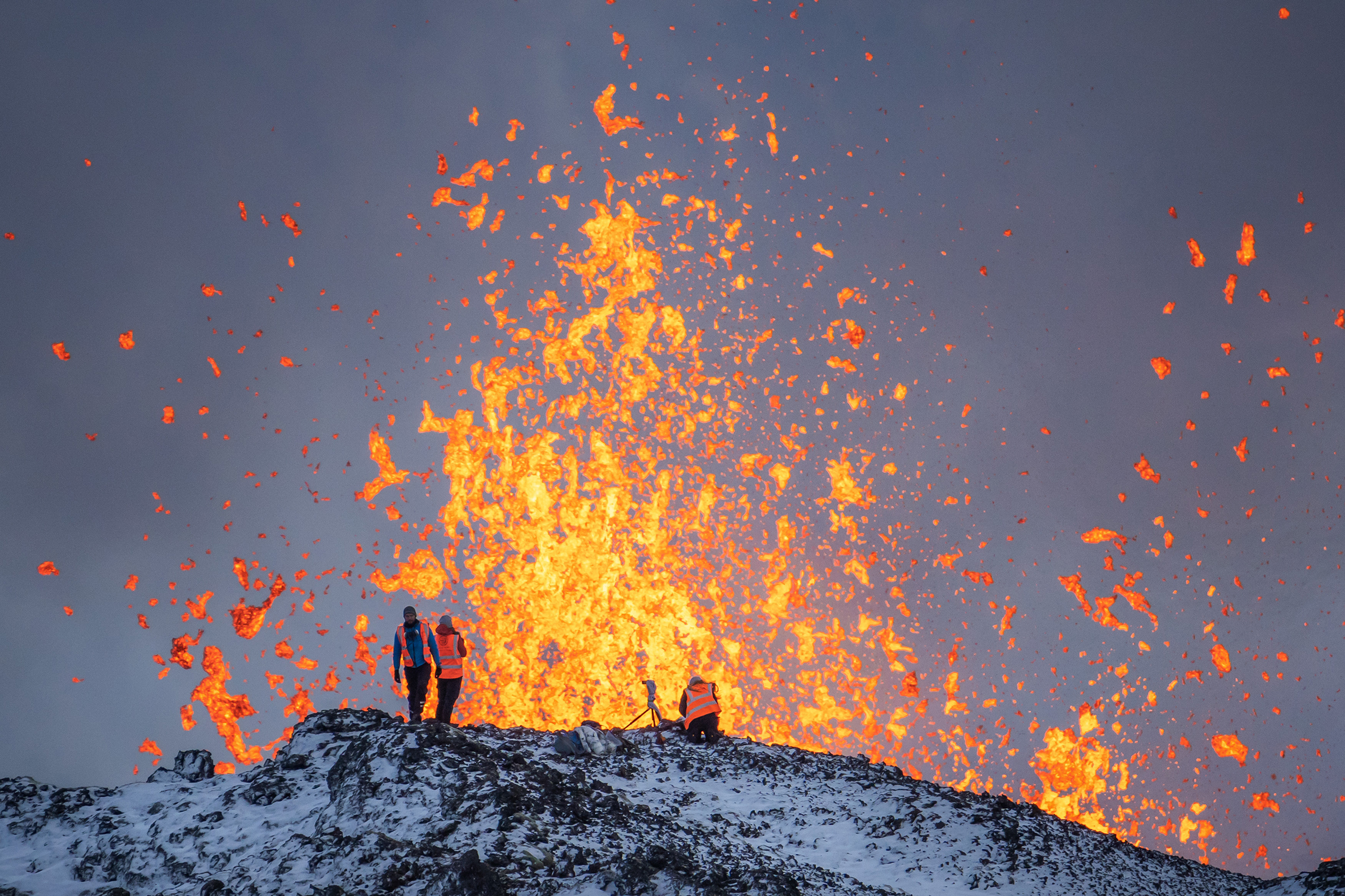 Three people stand on a volcanic landscape as lava erupts dramatically against a dark sky, surrounded by glowing orange splashes