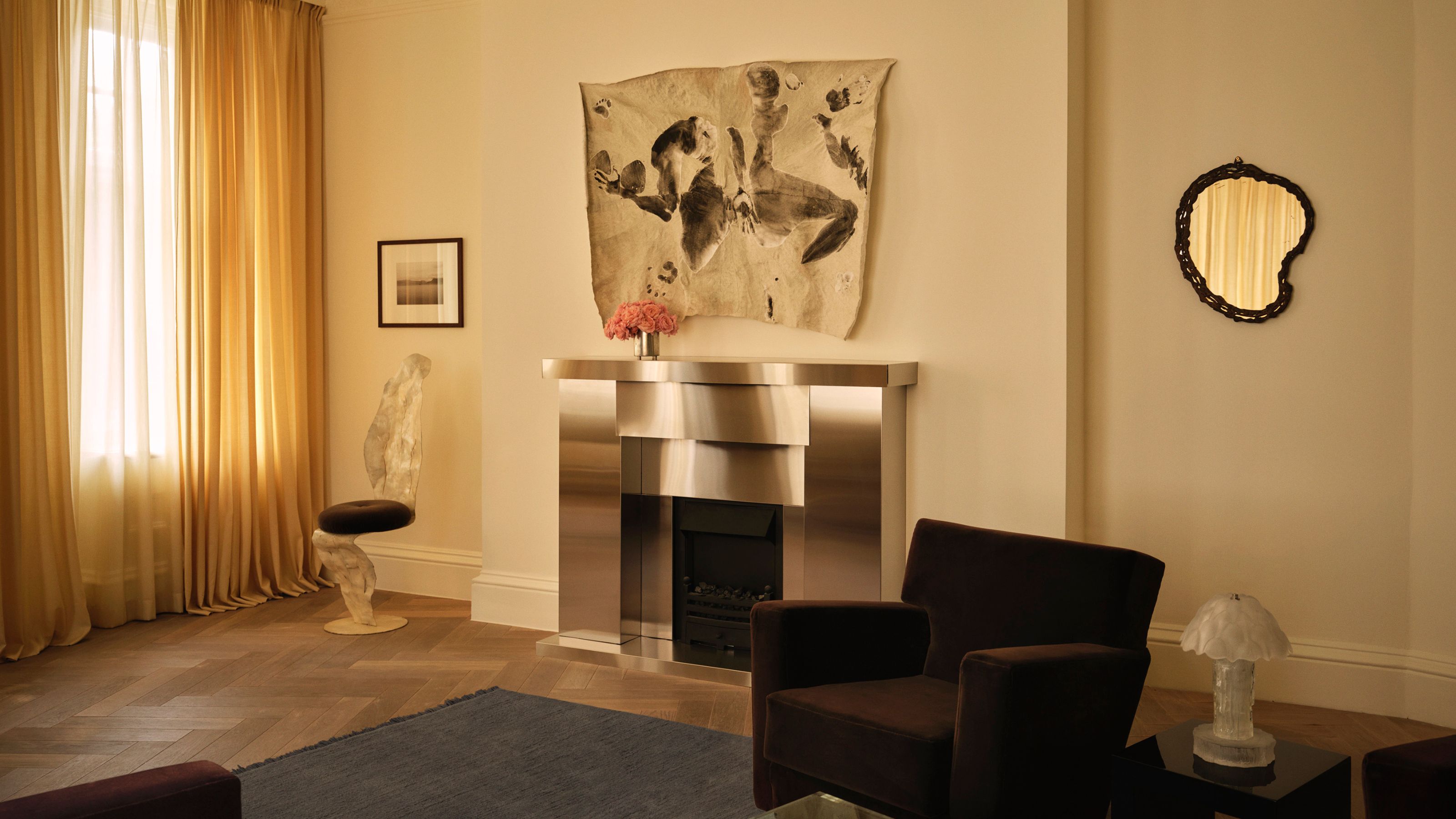 Nordic Knots London showroom with cream walls, a shiny metallic fireplace, wall mirror, tapestry over the fireplace, brown armchair, blue rug, timber floors and yellow curtains with light flowing through them