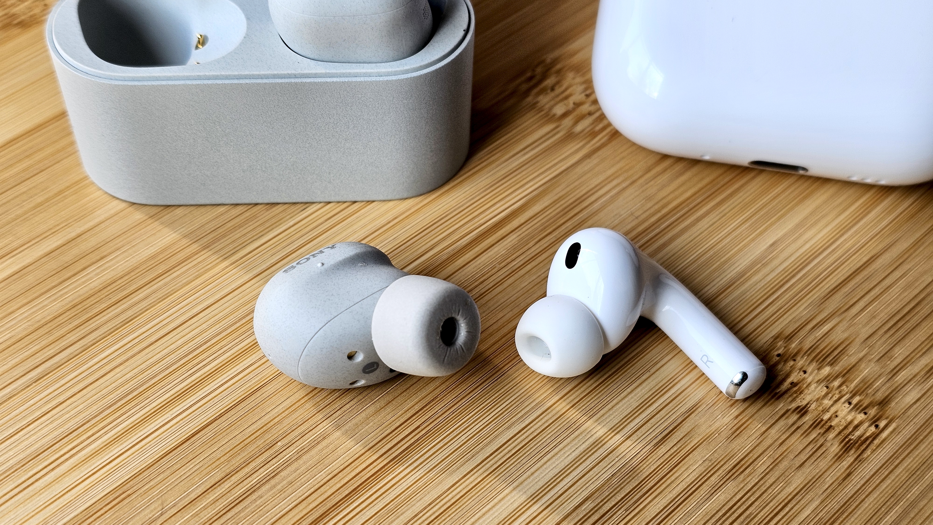 Sony WF-1000XM6 earbuds next to the Apple AirPods Pro 3, on a wooden table, in a versus-style format