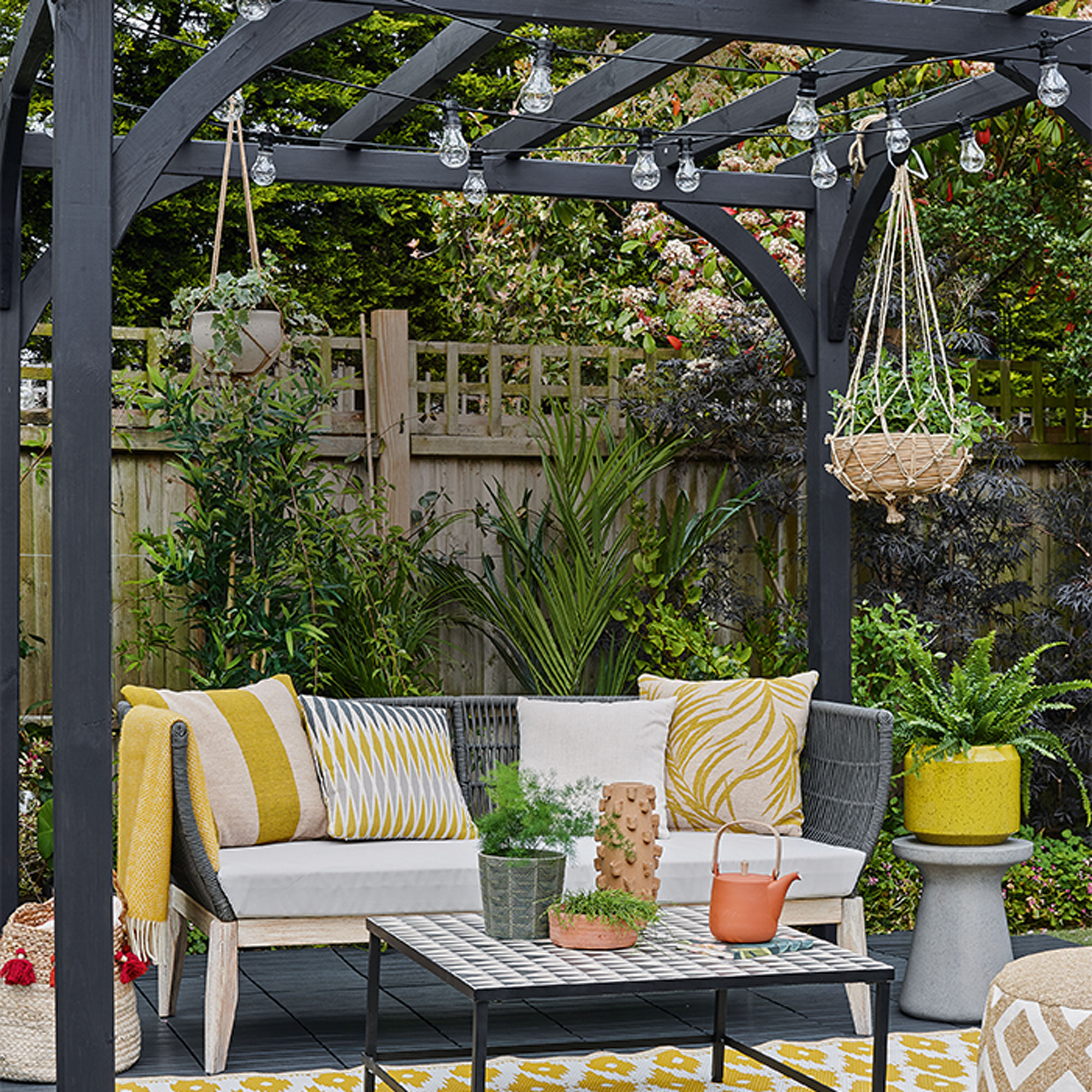 Black painted wooden pergola with string lights, hanging basket planter and garden bench with yellow and white cushions