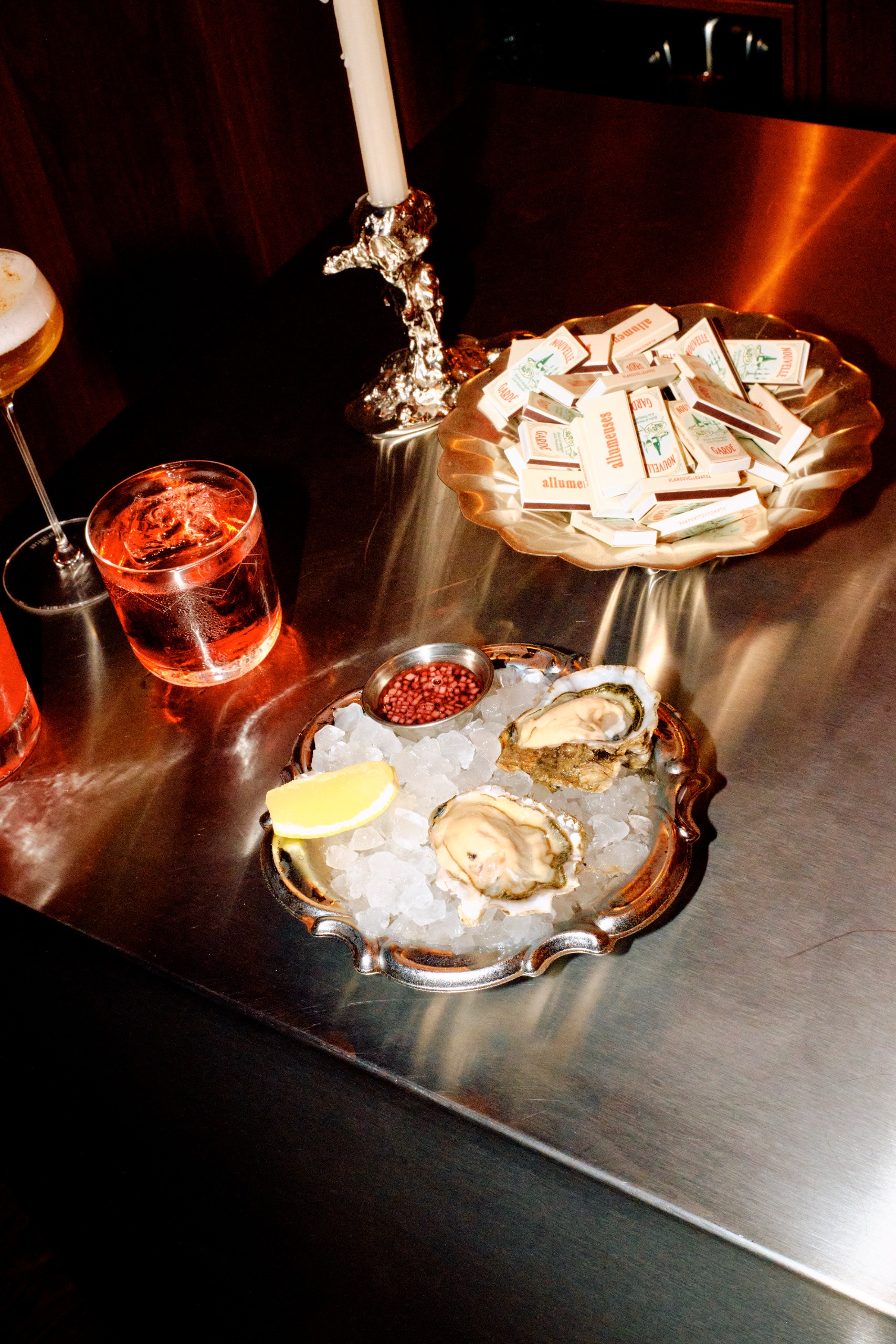 A flashed-out matte chrome countertop topped by silver bowls filled with oysters on the rocks and cocktails, alongside silver candelabra.