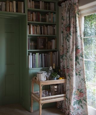 Country cottage style corner of a living room, painted green walls and woven, naturally textured drinks trolley, large floor to ceiling window with floral curtains, large bookshelf with books on wall and above doorway