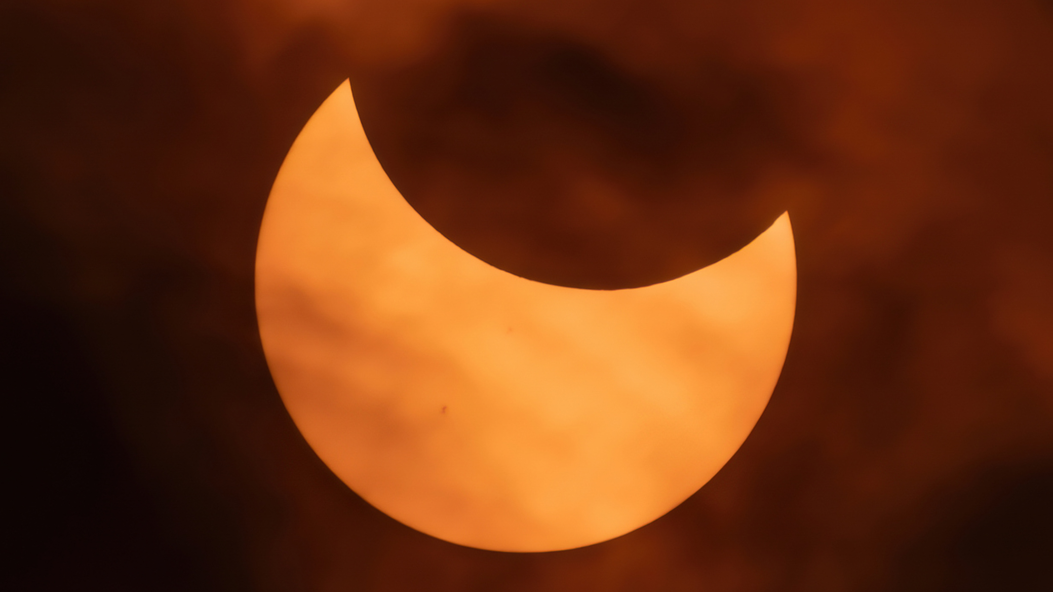 An image of the orange solar disk being partially eclipsed by the black lunar disk, with some clouds.