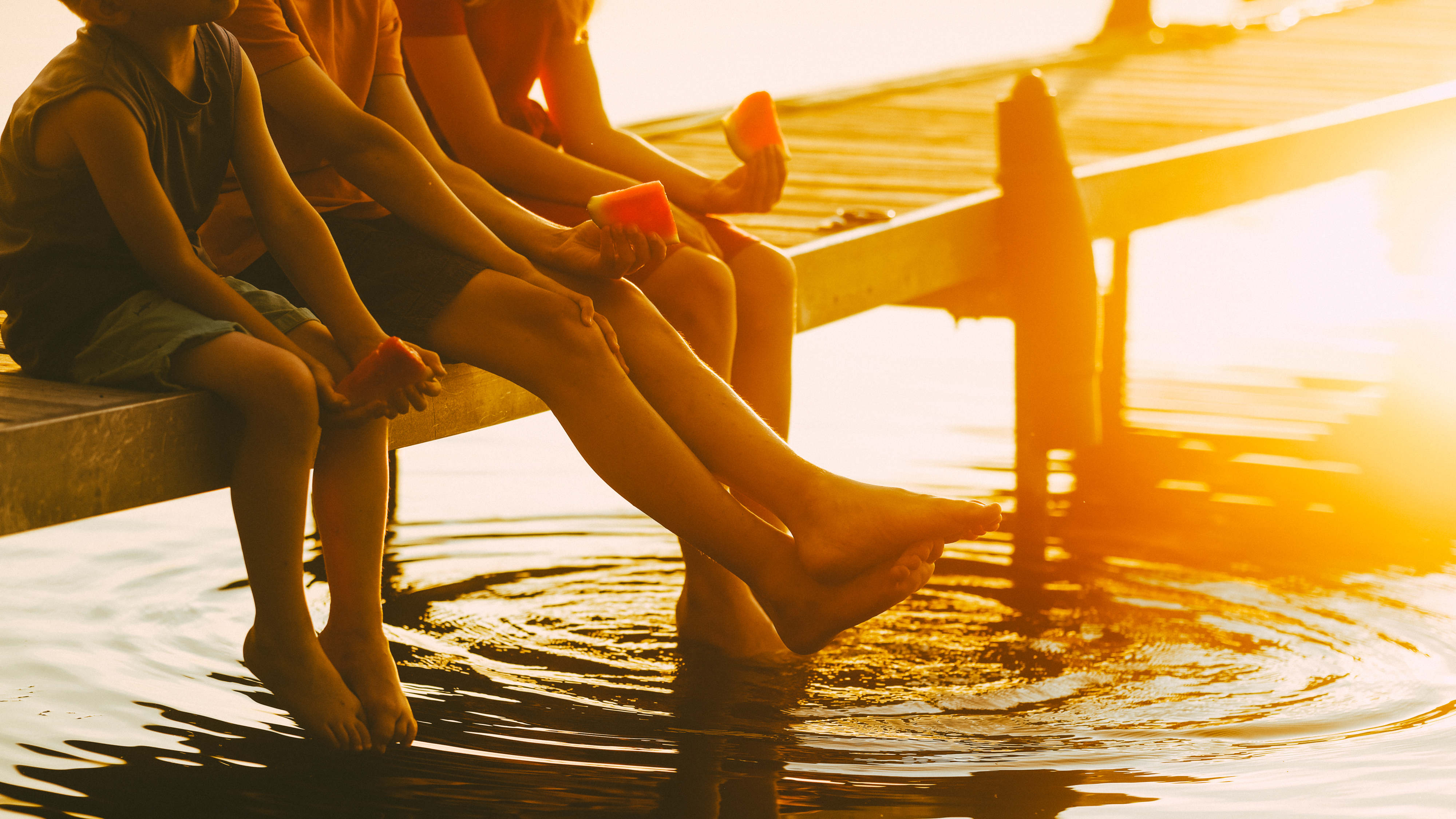 three children holding watermelon with their feet hanging off a dock with the summer sun shining off the water