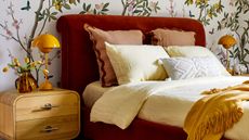 Image of a bedroom with a white wallpaper that has branches and green leaves. The bed frame is a dark red velvet and the bedding is a combination of yellows and reds.