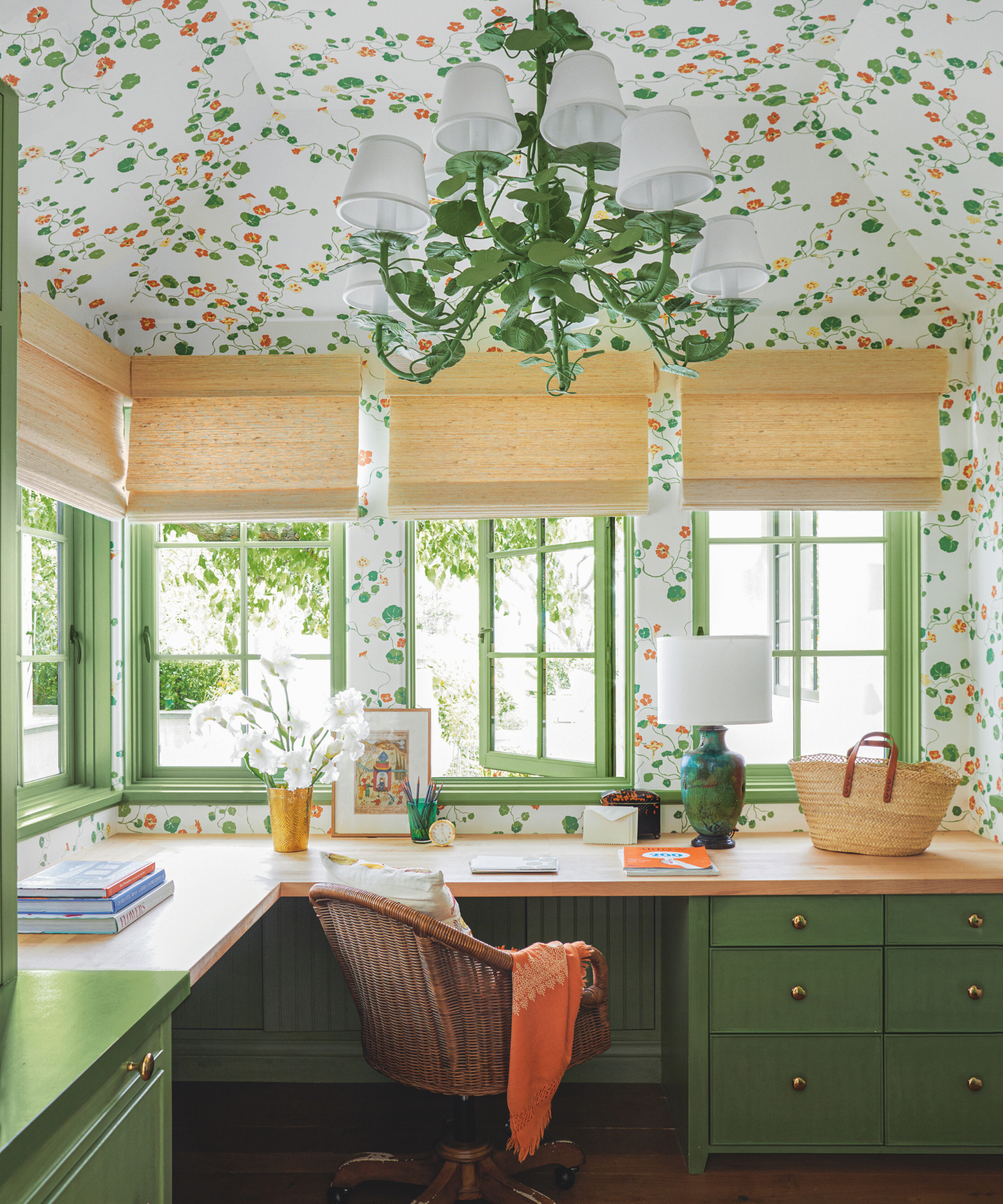 a home office with a built in countertop with green painted drawers, decor on the countertop surface, windows with blinds in front of them, and wallpaper in a floral pattern that continues up to the ceiling