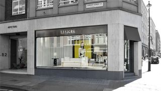 Exterior view of Christian Liaigre’s Conduit Street showroom. The window display features a light coloured sofa, lamps, a side table and a large image of a woman against a yellow background