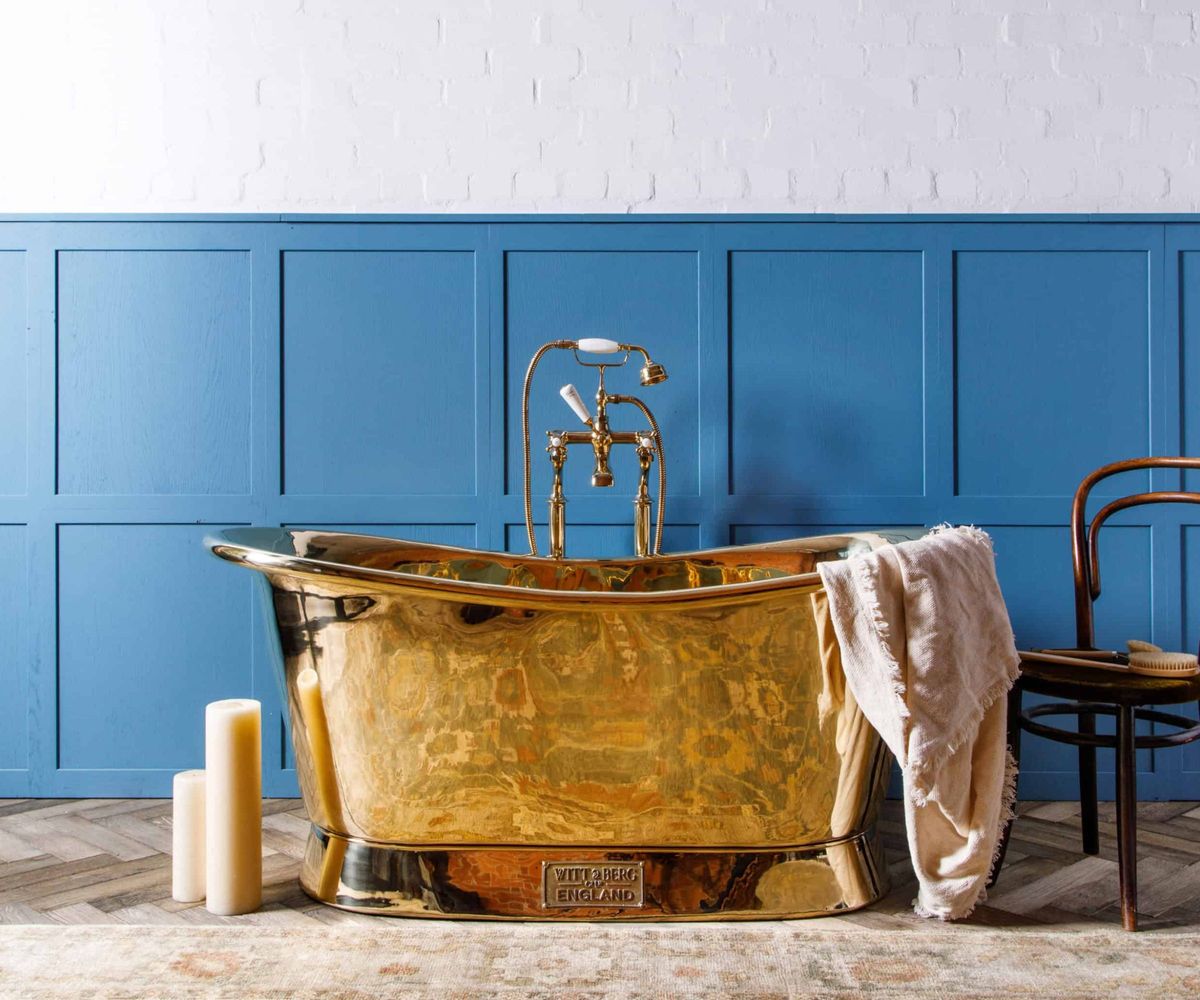 A brass standing bath with old fashioned brass taps set against a blue panelled wall 