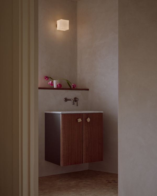 Image of a modern bathroom sink area as seen through a doorway. The walls are a beige limewash, and there is a floating wooden cabinet with white ceramic knobs.
