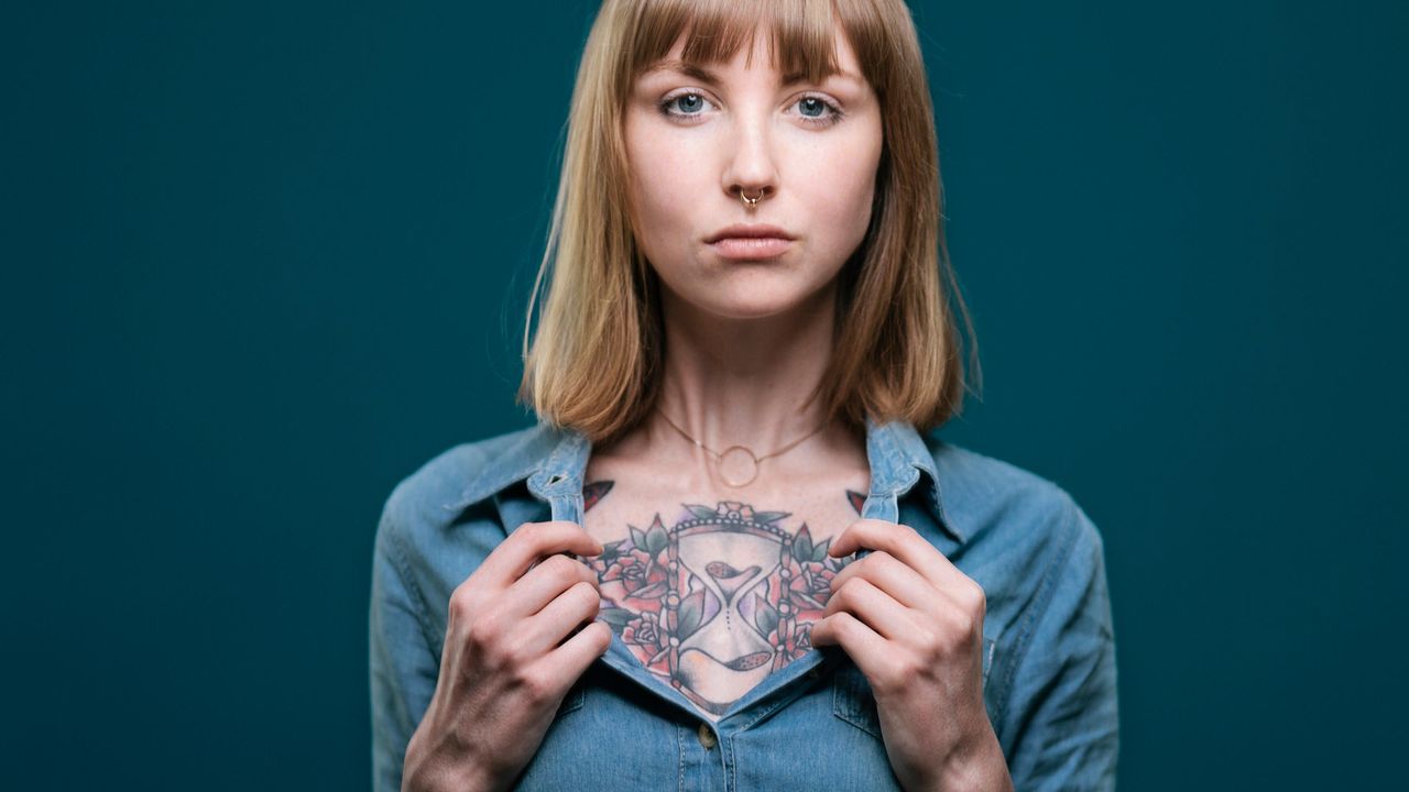 A woman opens the neck of her conservative blouse, revealing a large tattoo covering her chest.