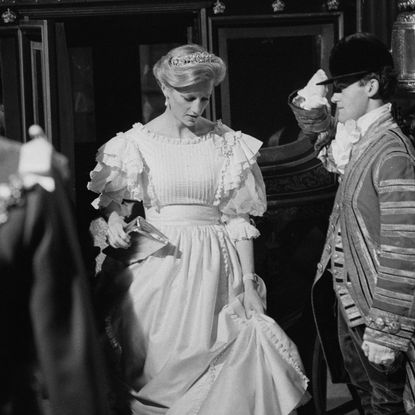 A black and white photo of Princess Diana in a long white dress and tiara with a footman saluting her