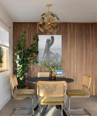 A dining room with a reclaimed wooden accent wall with a black and white bird print, a gold hanging light, a black circular dining table with a plant on top of it and four wooden rattan chairs around it