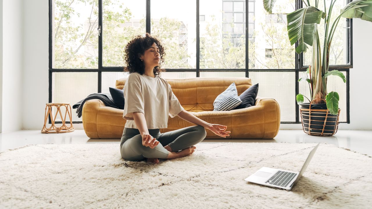 Woman sits cross legged on white rug in front of a couch. A laptop is open in front of her