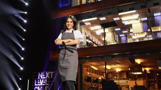 Contestant Gurpreet in a white top and grey apron in front of the kitchen in Next Level Chef.