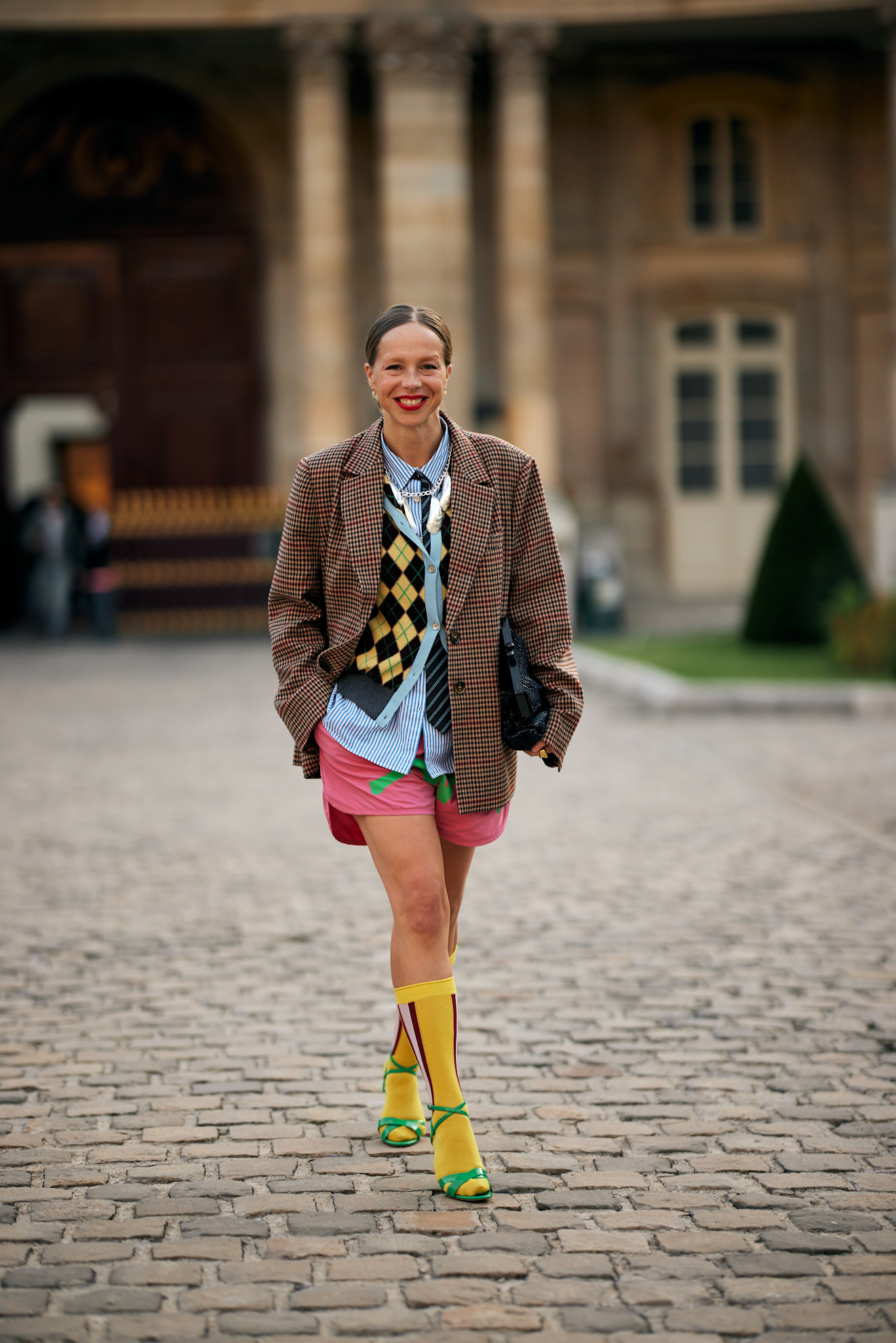 Chloe King wearing a black and yellow argyle sweater, blue button-down shirt, plaid blazer, pink shorts, yellow socks, and green heels at Paris Fashion Week