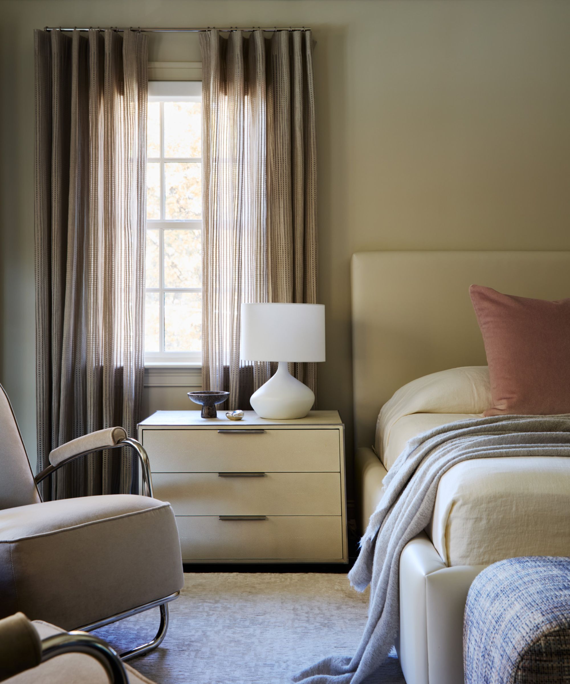 A bedroom with neutral walls, taupe curtains, and a warm white bed headboard with layered cozy bedding.