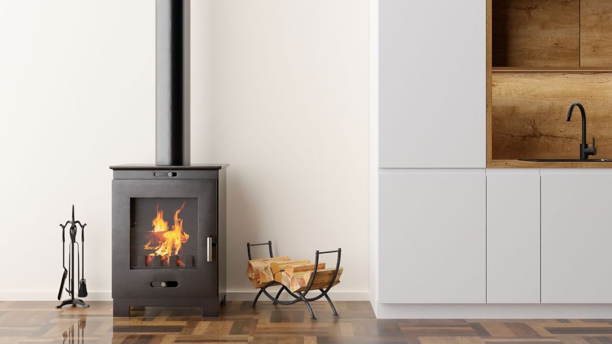 A sleek black wood-burning stove with a visible flame is positioned in a kitchen on a polished wood floor. Nearby are fireplace tools and a log holder filled with wood. Modern white kitchen cabinetry and wood accents complete the minimalist design.