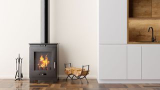 A sleek black wood-burning stove with a visible flame is positioned in a kitchen on a polished wood floor. Nearby are fireplace tools and a log holder filled with wood. Modern white kitchen cabinetry and wood accents complete the minimalist design.