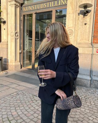 Danish female fashion influencer Sidsel Alling poses on a sidewalk in Copenhagen, Denmark, holding a glass of wine wearing a black blazer, white tee, a leopard-print mini duffel bag, and black jeans