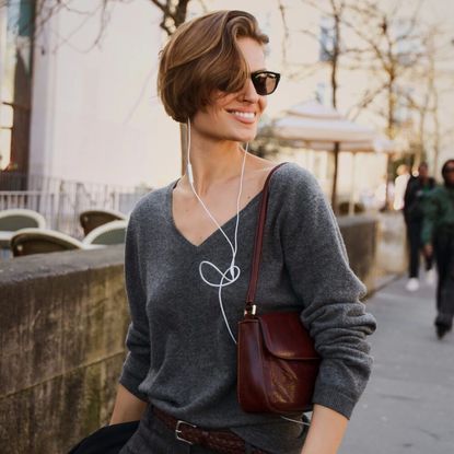 Woman poses while wearing gray sweater, black pants, and brown handbag