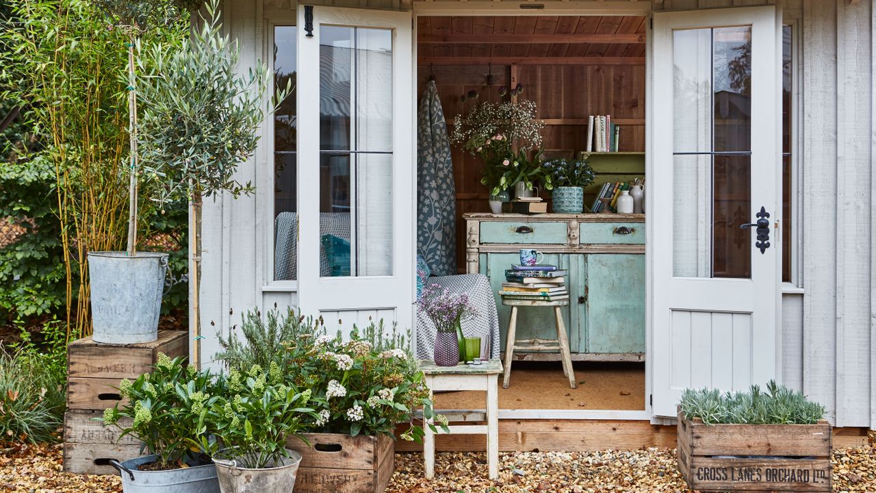 Storage ideas for sheds demonstrated with a shabby chic blue painted workstation inside a pale gray shed with potted plants outside on a gravel area.